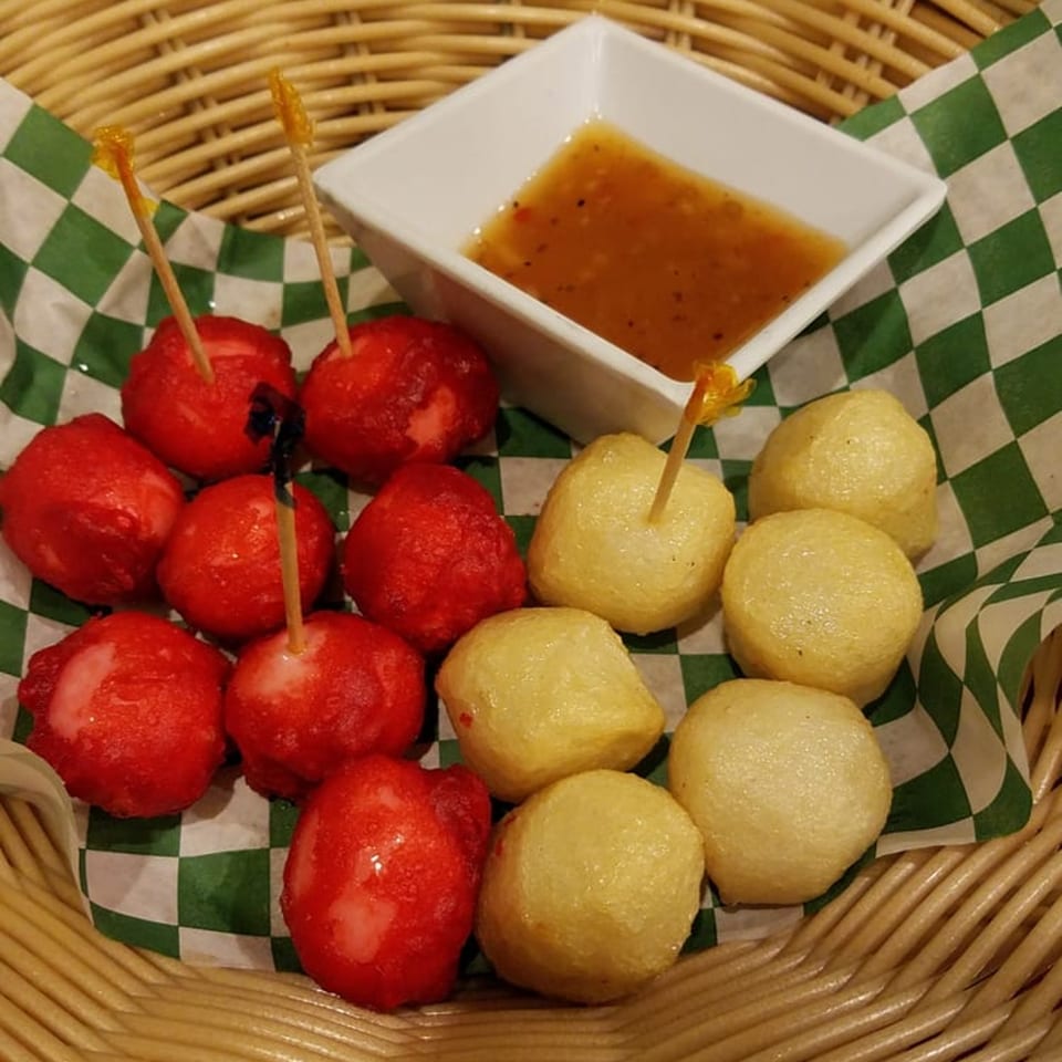 Red and yellow Filipino fish balls with toothpicks and a dipping sauce in a woven basket.